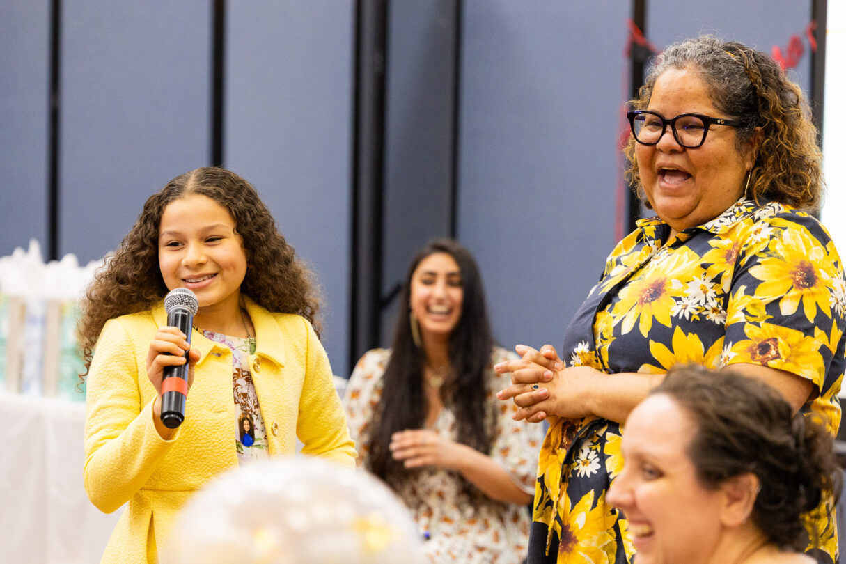 A young girl in a yellow sweater speaks into a microphone. Three women wearing yellow outfits laugh.