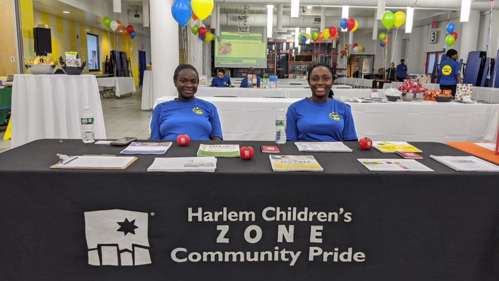 Two women sit and smile at their table to distribute information at a community fair.