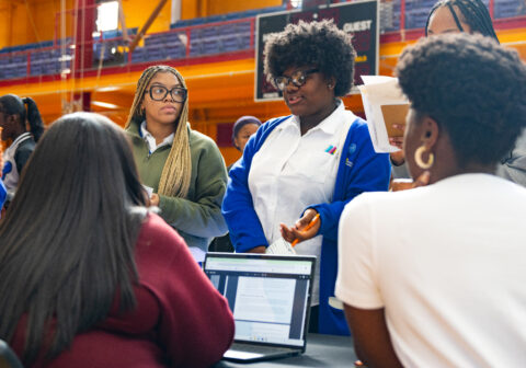Teenager in white shirt talks to woman in red shirt sitting at table with laptop.