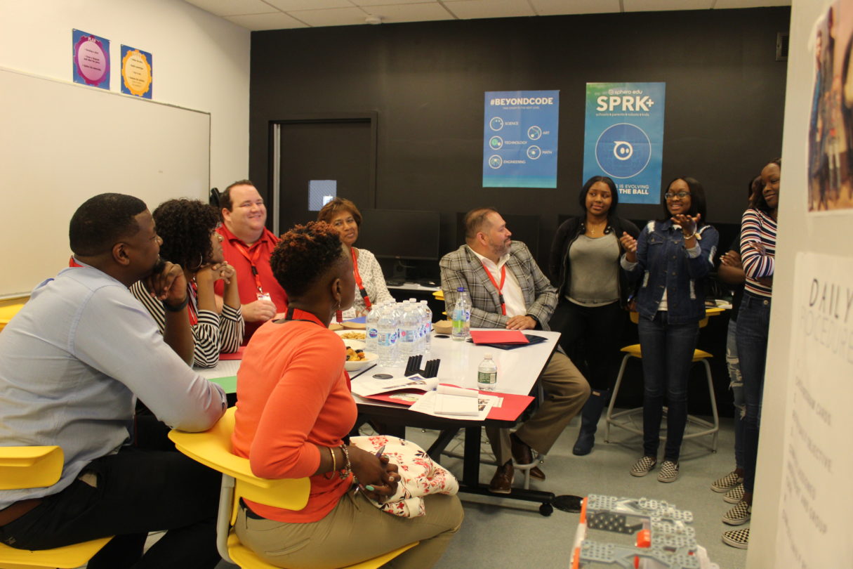A group of people listens to teenage girls giving a presentation in a classroom.