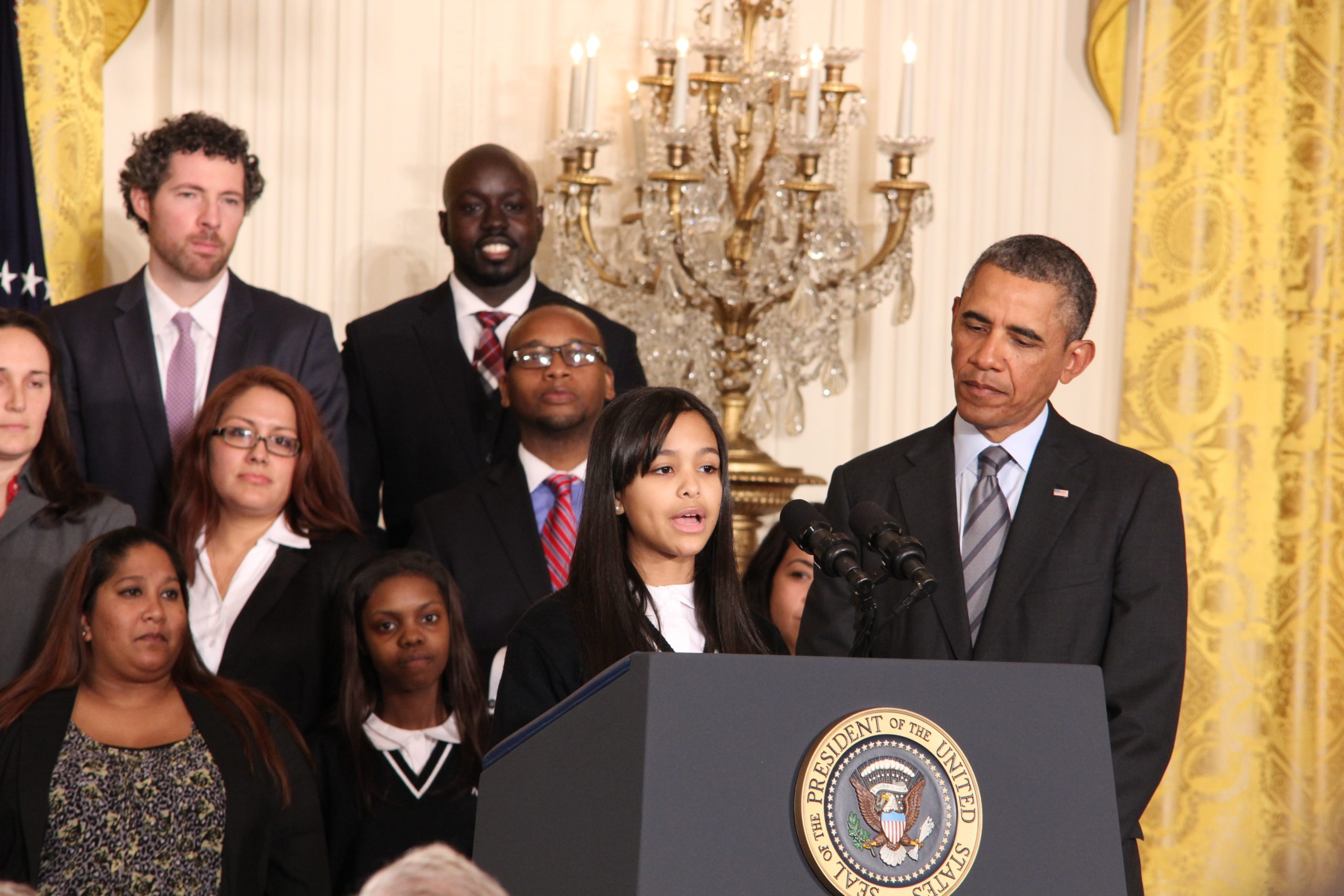 A young girl speaks at a podium at the White House. President Barack Obama, her classmates, and school leaders look on