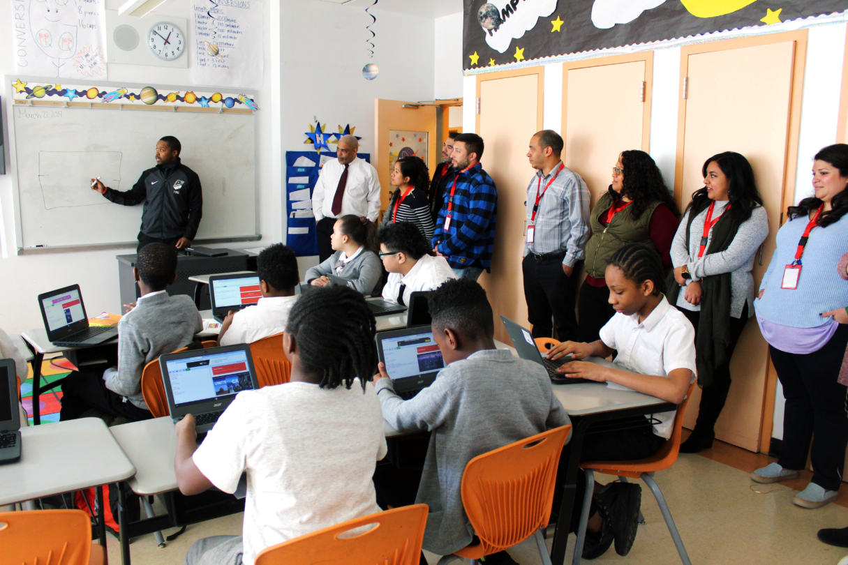 Practitioners Institute | A group of people with lanyards stand across a wall and observe an elementary class. A teacher points at the board as children read on laptops.