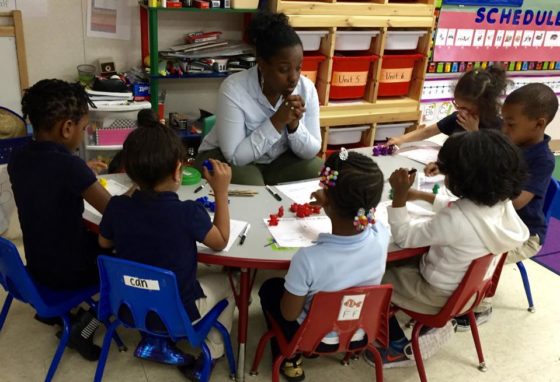 Teacher sits a table surrounded by young students