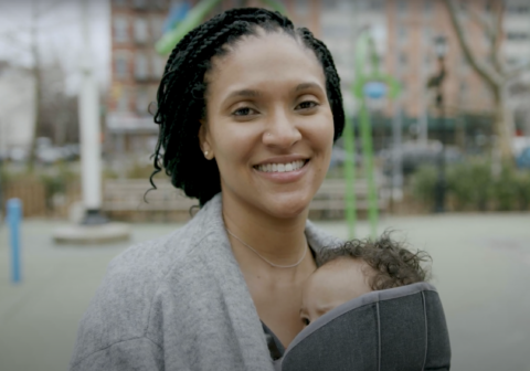 Reyna Bonaparte in a grey sweater holds her baby and smiles.