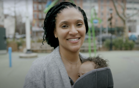 Reyna Bonaparte in a grey sweater holds her baby and smiles.