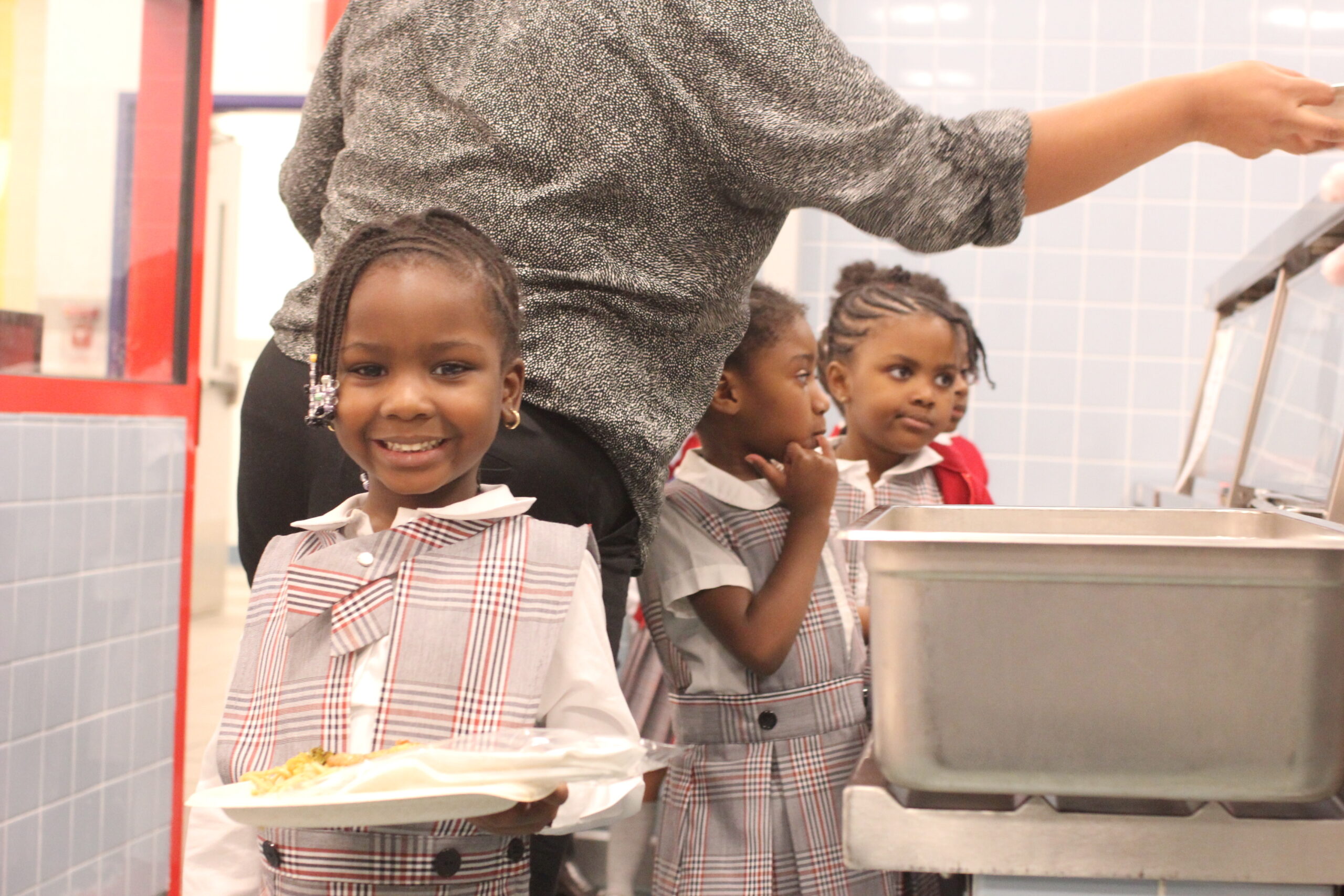 A young girl in a school uniform holds a tray of food in a cafeteria.