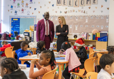 Harlem Children's Zone CEO Kwame Owusu-Kesse, wearing a maroon suit, standing next to Tracy Smith, CBS Sunday Morning correspondent, standing in classroom with young students.