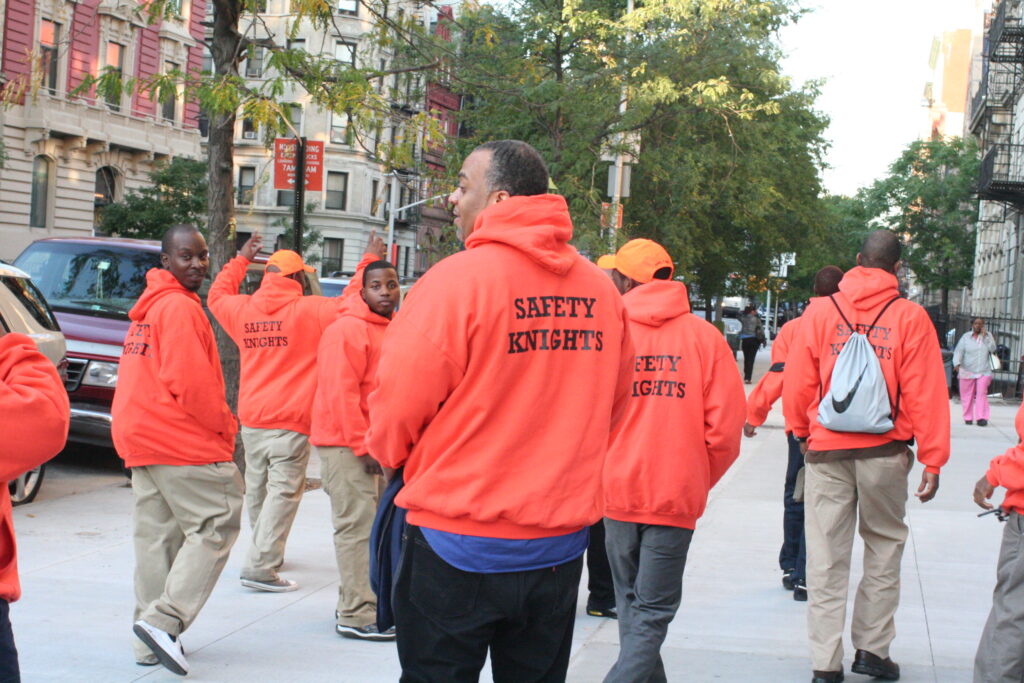 A group of people wearing bright orange sweatshirts with the text "Safety Knights" on them walk down the street.