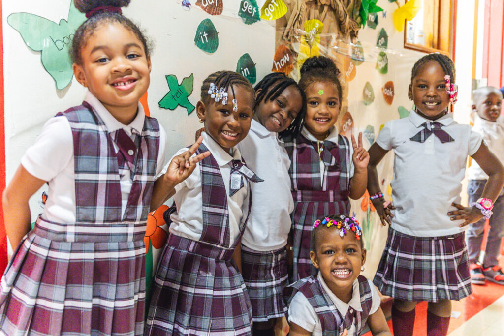 Children in burgundy-plaid uniforms smile and pose in a school hallway.