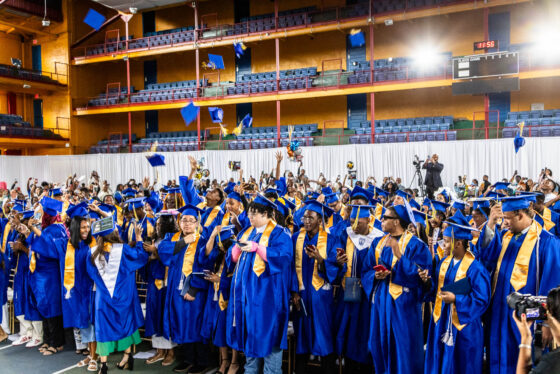 Teenagers in blue-and-gold graduation gowns throw their caps into the air to celebrate their graduation.