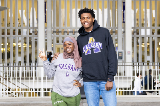 Two students. Brandon Sydnor, a male student in a black sweatshirt with the text "U-Albany" on it, and a female student wearing a headscarf and a gray sweatshirt with purple "U-Albany" lettering on it.