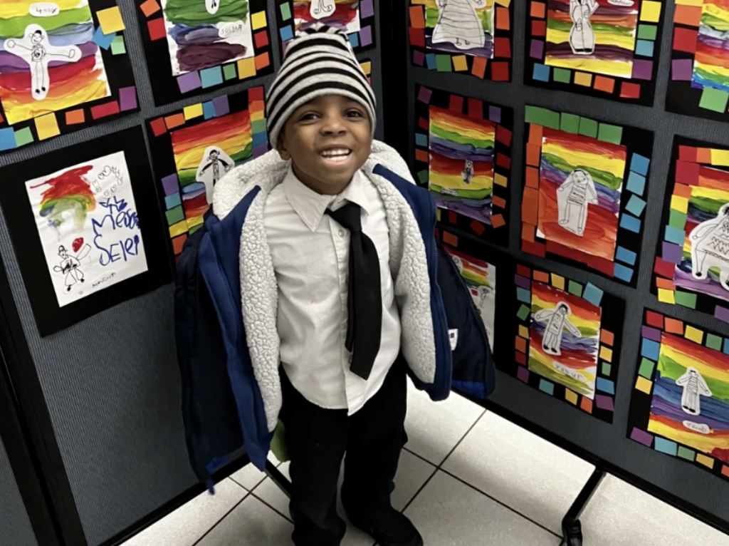 Reginald, a young student wearing a school uniform, stands in front of class artwork.