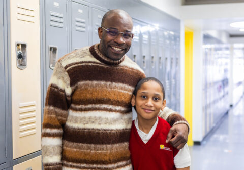 Man in striped sweater and child in red vest stand near lockers.