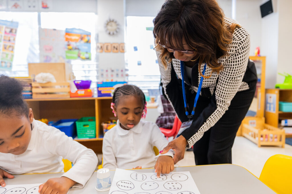 A woman stands next to a young female student in a classroom