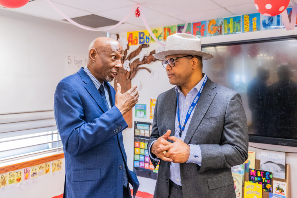 A man in a blue suit talks with man in a gray suit inside a classroom