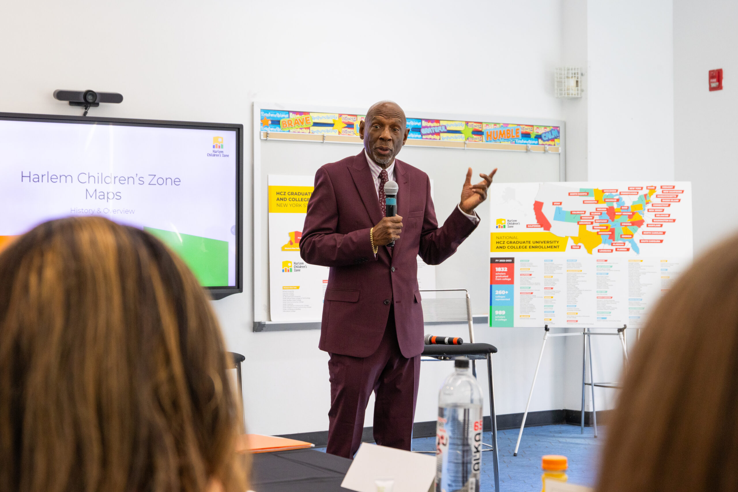 A man in a burgundy suit gives a presentation in front of a classroom.