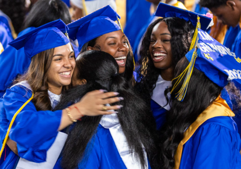 A group of female students in blue caps and gowns smile and hug each other.