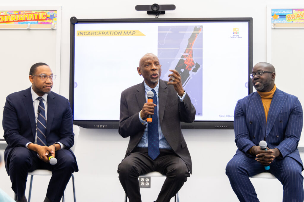Geoffrey Canada seated beside two male professionals as he speaks into a microphone