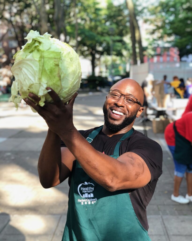 Man smiling and wearing an apron holds up a large head of lettuce with two hands.