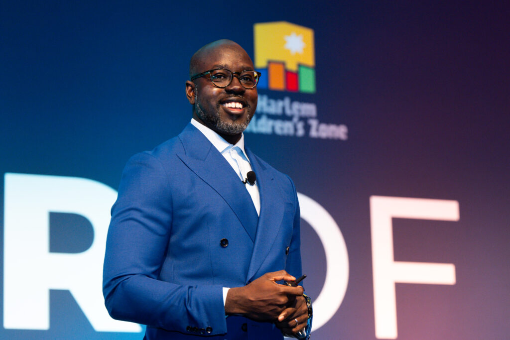 CEO of Harlem Children's Zone Kwame, smiles and looks off into the distance while standing on stage in a dark blue suit.
